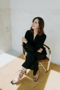 Jill van den Bosch op een stoel, ze kijkt naar de zijkant en draagt een zwart pak